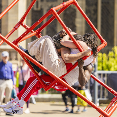 Three people stand in a line in the sun, with linked arms, in front of a red scaffolding cube. The first two are a Black man wearing a silver top, silver hat, red neckerchief and red trousers, with his thumbs outstretched, and a white woman with curly black hair wearing a black bucket hat, white top, silver waistcoat with white tassels, and red shorts. She is smiling. The third man has his back facing the audience, and one hand outstretched. He is a brown man wearing a black hat, black leather waistcoat and red tartan trousers.
