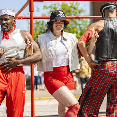 Three people stand in a line in the sun, with linked arms, in front of a red scaffolding cube. The first two are a Black man wearing a silver top, silver hat, red neckerchief and red trousers, with his thumbs outstretched, and a white woman with curly black hair wearing a black bucket hat, white top, silver waistcoat with white tassels, and red shorts. She is smiling. The third man has his back facing the audience, and one hand outstretched. He is a brown man wearing a black hat, black leather waistcoat and red tartan trousers.