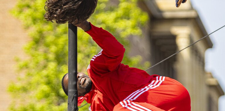 Two people are suspended horizontally from a black rod, holding on with their arms. Above is a white woman with mid-length curly brown hair, wearing a white tank top and beige trousers, hanging upside down with her legs extended forward at a right angle. Below her is a Black man in a red tracksuit and white sneakers, gripping the rod with bent arms spread wide and his legs curled toward his body.