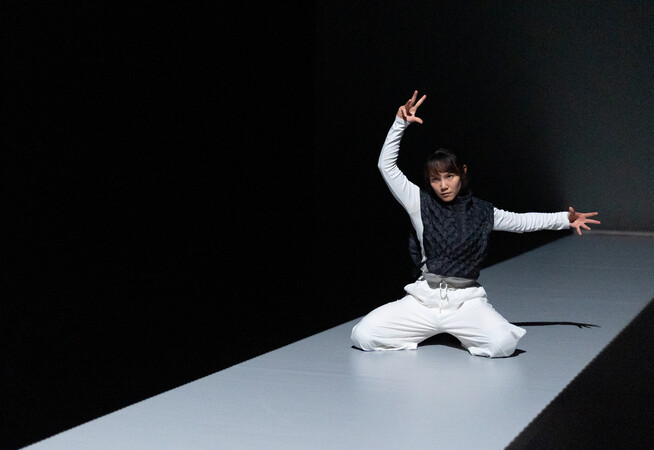A dancer kneels on a narrow, illuminated stage, wearing white pants and a dark textured vest, striking a dramatic pose with one arm raised and the other extended outward.