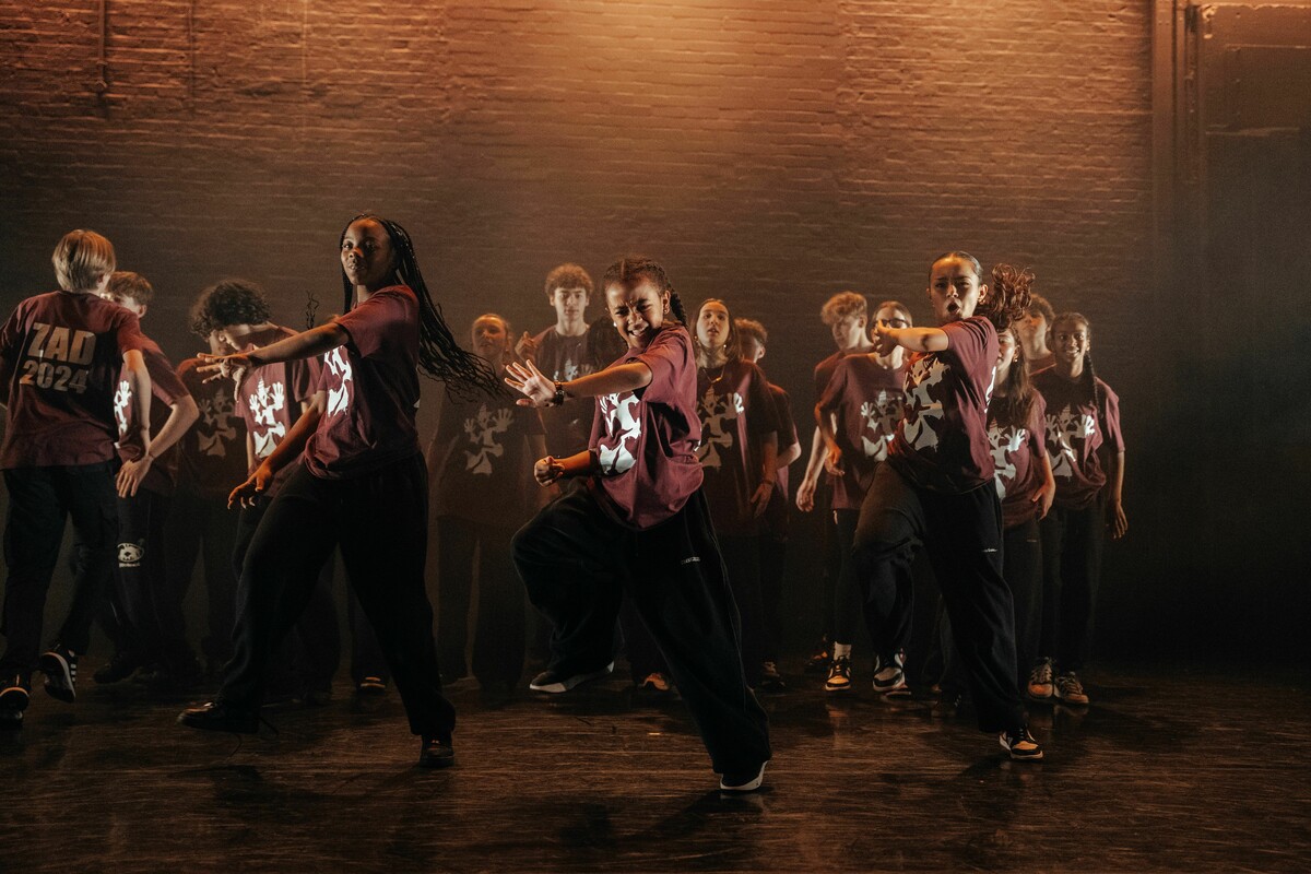 A group of children in matching burgundy T-shirts with white logos, black trousers, and sneakers dancing on stage.