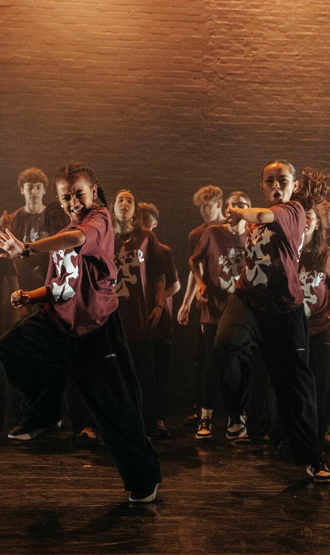 A group of children in matching burgundy T-shirts with white logos, black trousers, and sneakers dancing on stage.