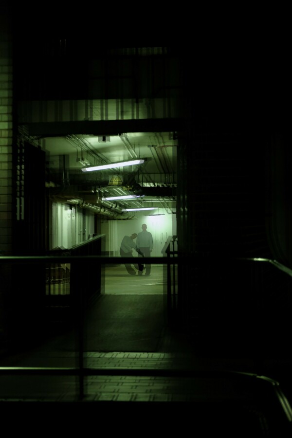 Green-tinted, slightly blurred image of a dark building entrance, showing a person in the distance, appearing twice, standing still and leaning left, as if caught in motion.