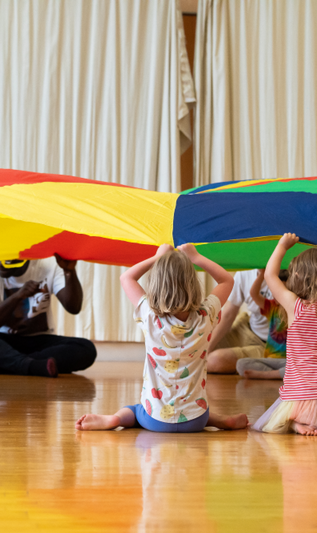 A group of children sitting in a circle on the floor holding up a parachute.