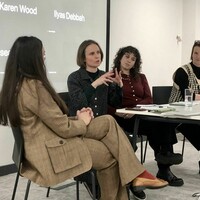 Four people sit in a panel discussion, with Christina, second from the left, speaking while the others listen. A projection screen with participant names is visible behind them.