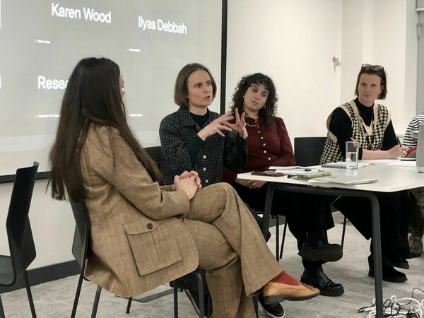 Four people sit in a panel discussion, with Christina, second from the left, speaking while the others listen. A projection screen with participant names is visible behind them.