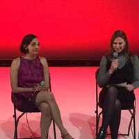 Two people sit on chairs onstage against a red backdrop, with Suzanne on the right. The person on the left, holding a microphone in her lap, wears a sleeveless purple dress. The person on the right holds a microphone and reads from a paper, dressed in a grey turtleneck with a black dress layered over it. A bottle of water sits on the floor beside them.