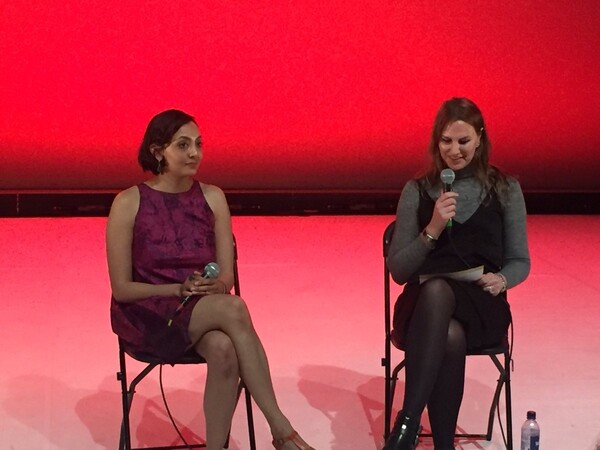 Two people sit on chairs onstage against a red backdrop, with Suzanne on the right. The person on the left, holding a microphone in her lap, wears a sleeveless purple dress. The person on the right holds a microphone and reads from a paper, dressed in a grey turtleneck with a black dress layered over it. A bottle of water sits on the floor beside them.