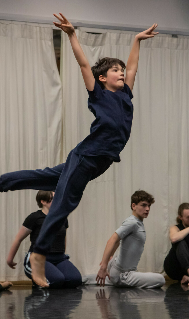 A young boy mid-jump, surrounded by male-identifying young dancers of various ages kneeling on the ground and looking in different directions