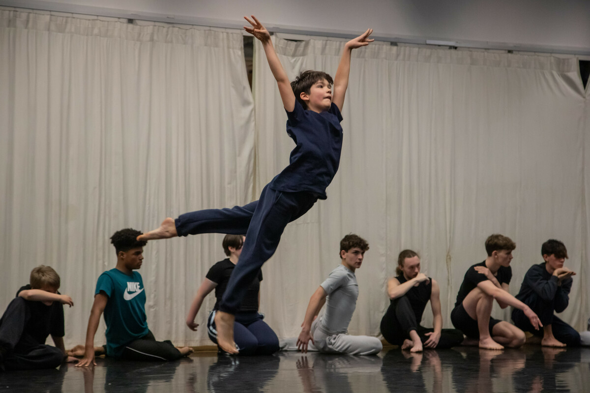 A young boy mid-jump, surrounded by male-identifying young dancers of various ages kneeling on the ground and looking in different directions