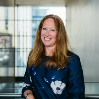 A woman with light red hair and a big smile looks at the camera. She is leaning on the metal railing and wearing a blue silk long sleeved top.