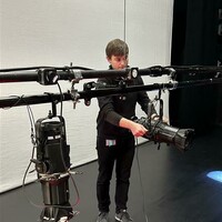 Luke on stage adjusting a reflector spotlight. He has short brown hair and is wearing a black long-sleeve top, black trousers, and grey sneakers.