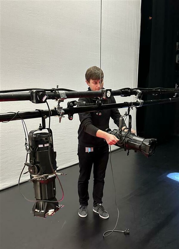 Luke on stage adjusting a reflector spotlight. He has short brown hair and is wearing a black long-sleeve top, black trousers, and grey sneakers.