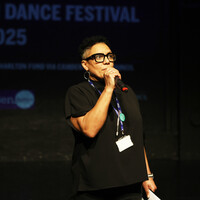 Maria on stage speaking into a microphone. She has short black curly hair, wears glasses with thick black frames, and is dressed in a black t-shirt, black trousers, and a blue lanyard.
