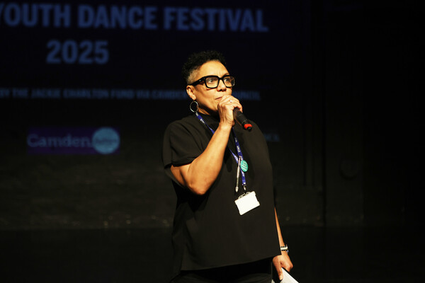 Maria on stage speaking into a microphone. She has short black curly hair, wears glasses with thick black frames, and is dressed in a black t-shirt, black trousers, and a blue lanyard.