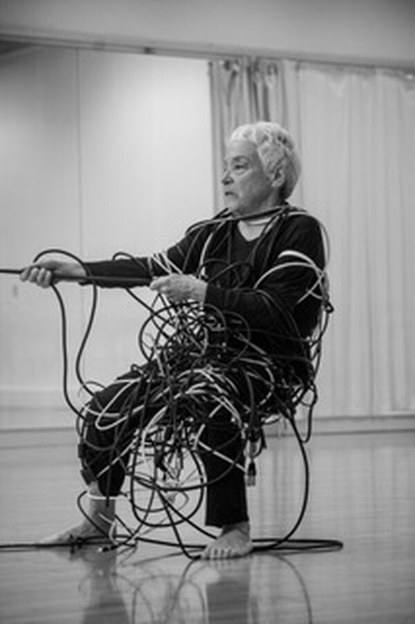 Black-and-white photo of an older adult sitting in a studio, wrapped in a large bundle of cables and holding one cable while looking to the side.
