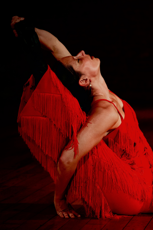 ChatGPT powiedział: A dancer in a red fringed outfit performs a deep back-arched floor pose on a dimly lit wooden stage.