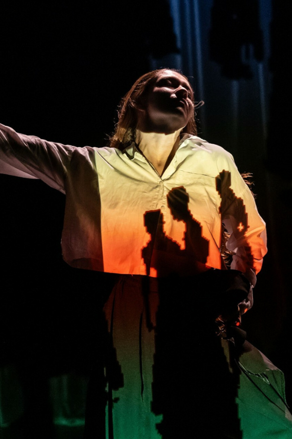 A person in a white shirt and black trousers raises one arm sideways in dim, warm light, with shadows across their shirt forming three silhouetted figures.