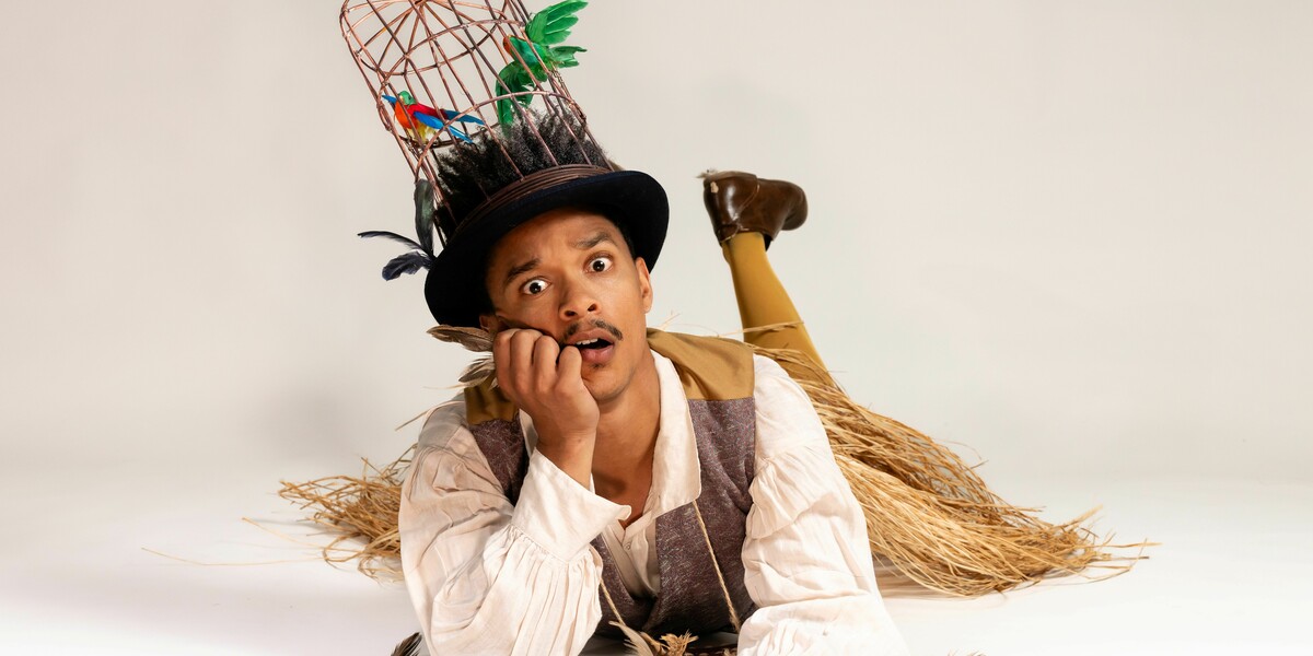 A person lies on the floor wearing a tall birdcage hat with artificial birds, holding a feather and looking surprised.