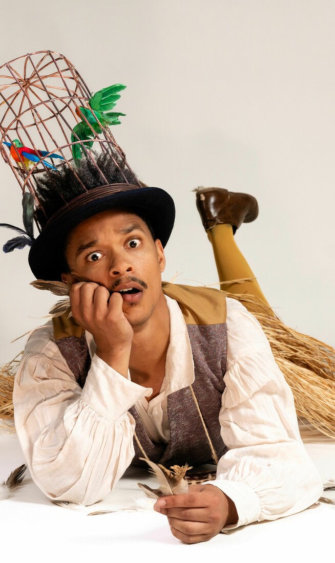 A person lies on the floor wearing a tall birdcage hat with artificial birds, holding a feather and looking surprised.