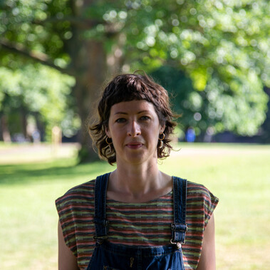 A close-up shot of a white woman in her 30s. She has short medium brown hair with a short fringe and is smiling against a background of trees in a park.