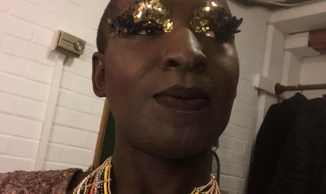 A photo showing Ebony Rose Dark, a black cabaret and dance performance artist with gold glitter around my eyes and dark lipstick on. They have contemporary jewellery around their neck which is multicoloured, gold, red, yellow. They are wearing a gold sequin glitter dress.