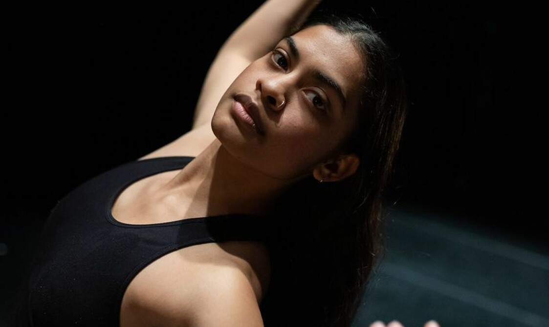 A close-up image of Chandenie, who is a young person of Indian heritage. They are standing on a stage with their arms up in the air looking into the camera while leaning slightly back. They are wearing a black crop-top and black trousers. Their long dark hair is worn down and they are wearing golden ear hoops and nose ring.
