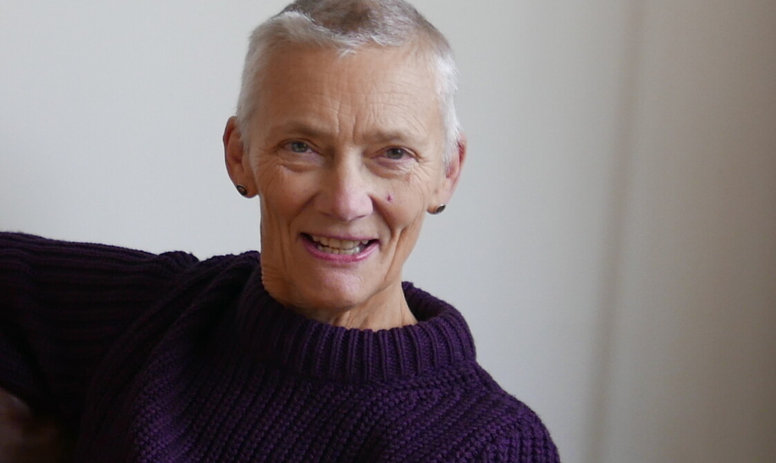A colour image of Emilyn Claid, a white woman at the age of 69. She is looking directly to camera and smiling. She has short-cropped silver hair and wears a purple jumper. Her right arm is thrown over the back of a chair, and her body is leaning slightly to her left.