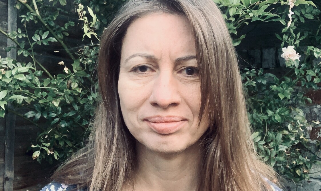 A colour photograph headshot of Holly, a white woman in her late 40’s with shoulder length brown hair, brown eyes and a soft gaze. Facing the camera, she wears a short sleeved blue patterned top. Behind her a rose bush adorned with white flowers, meanders in all direction over an ageing garden fence.