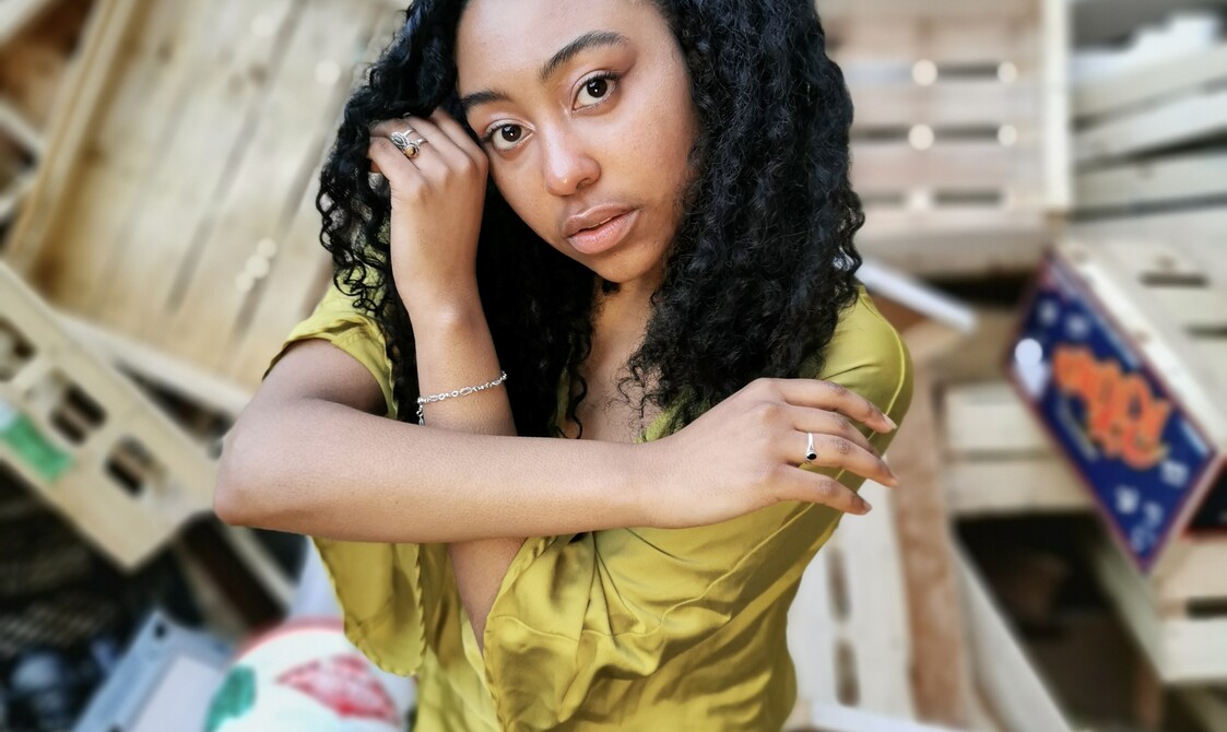 Paris wears a olive willed dress in front of a wooden garbage background. Her arms are twisted framing her face and she is front on to the camera with a tilted head.