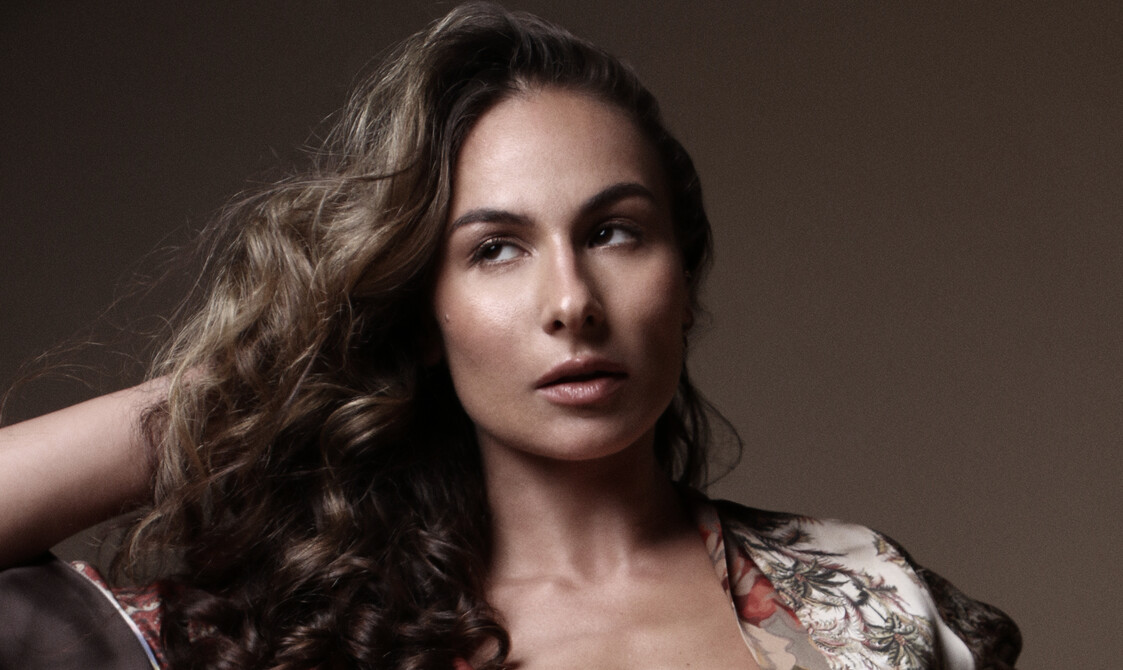 A colour photograph of the dance artist Stephanie Handjiiska, a Caucasian woman in the beginning of her 30s, on a soft brown background, with a direct photographic light cast on her. She looks up and right, her hair is in motion, and the sleeves of her brown kimono with white and orange flowers, fly up in the air and create interesting shapes in the wind.
