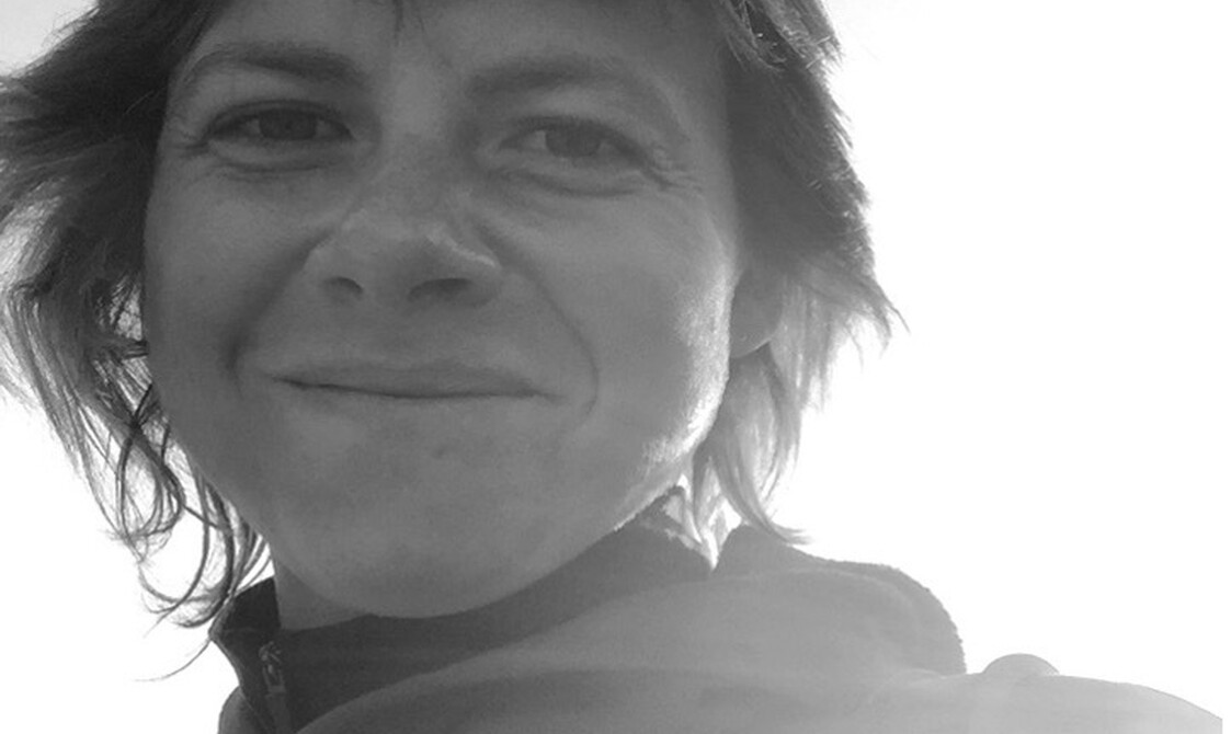 A black and white photo of Sophie Clements. a white woman in her mid 30s, Sunshines over her left shoulder. She is looking down with smile. Dark brown, short hair and wearing two jumpers.