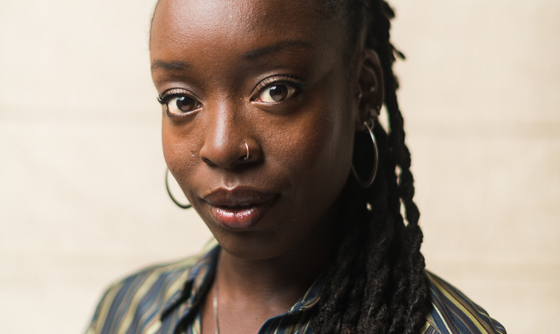 A colour photograph of Amarnah Ufuoma Cleopatra, a black woman in her late 20s. She has long twisted black dreadlocks, half up in a bun, half down hangin over one shoulder. She wears a striped shirt in green, brown and blue tones, a silver necklace with a green/grey/blue labradorite pendant, a slim silver nose ring and silver hooped earrings along with multiple studs dotted on different parts of her ear lobe. Her bright brown eyes are looking directly at the camera with a warm and present facial expression.
