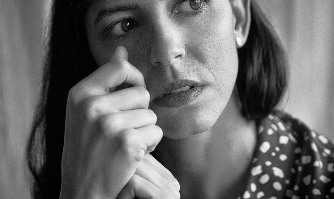 A black and white portrait of Stefania Pinato, a white woman in her early 30s. She has long black hair and a fringe, she is looking to her left side as she holds her hands in front of her right cheek. She is wearing a grey shirt with white uneven dots, and behind her there is white fabric as a backdrop.