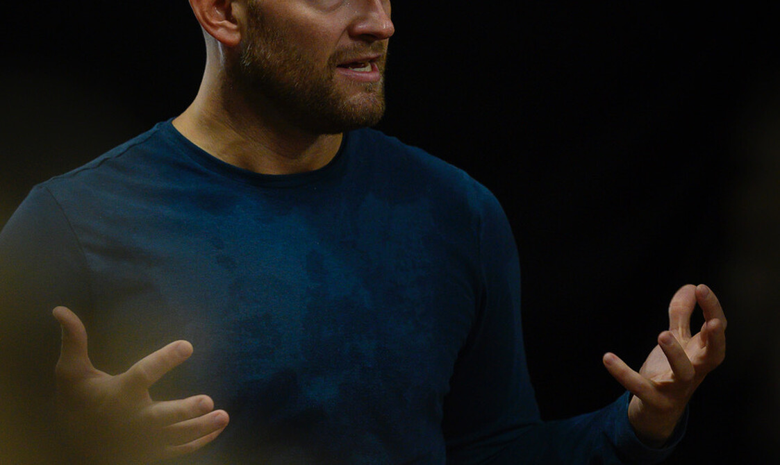 A photo of Hannes Langolf, a white man in his late 30s with a shaved head and a short ginger-blonde beard. He is wearing a sweaty, blue, long-sleeve shirt, standing in front of a black background. He is talking to a group of people, out of shot, his mouth open in conversation and his open hands held in front of his chest in an explanatory gesture.
