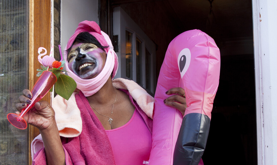 An older black woman stands in her doorway smiling as she takes a sip from a pink cocktail glass. She wears a bright pink unitard costume and bright pink flamingo rubber ring complete with yellow tights.