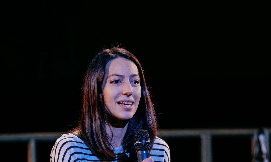 Adrienne Hart, a white woman in her late 30's, on a black stage with a gong circle and mic in the background. She has a microphone held up towards her face and is looking out. She has long dark hair and is wearing a stripy black and white top.