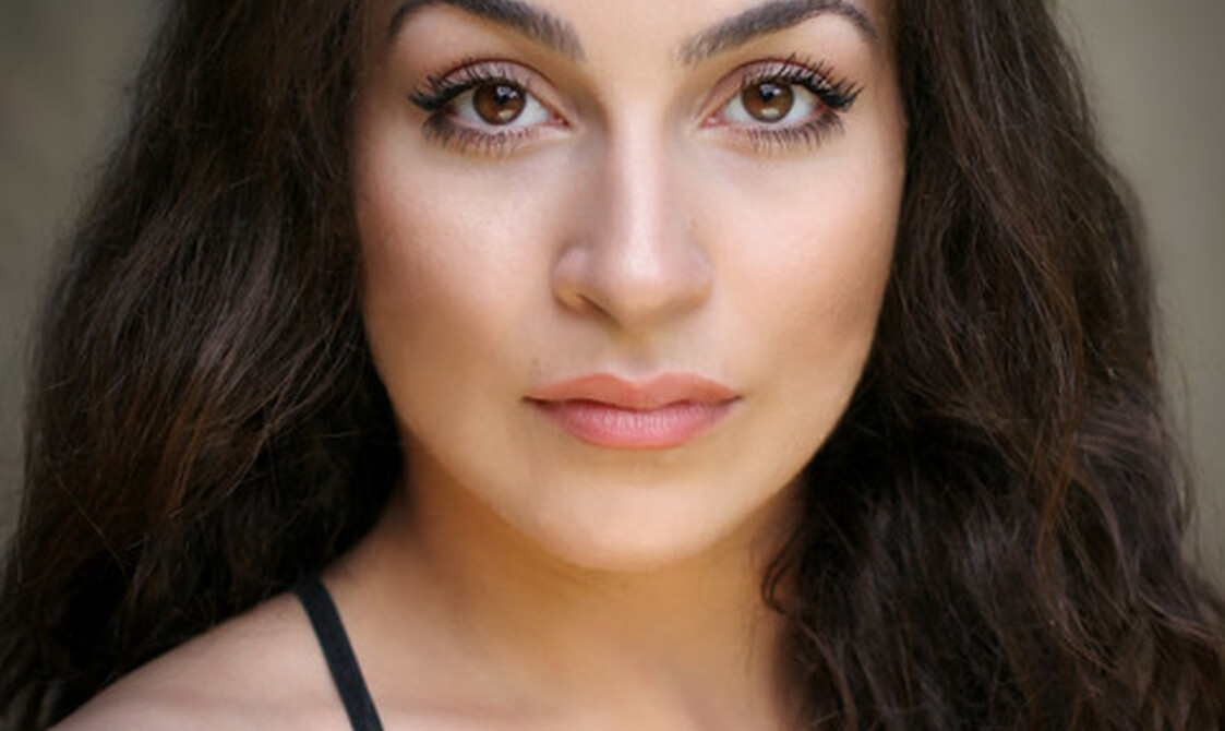 Woman with long brown wavy hair wearing a black strappy top looks into the camera.