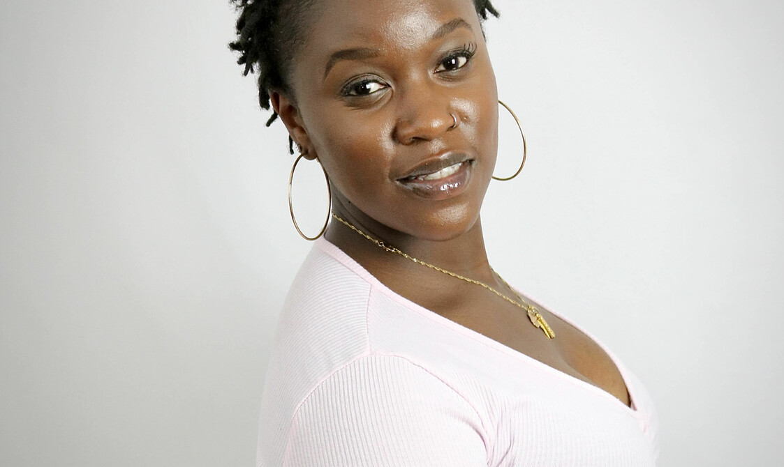 A photo of Sarah Lisney, a dark skinned lady of African heritage. She has short black loc’ed hair, dark brown eyes and is wearing a light pink long sleeved top. She is also wearing big gold hooped earring and a gold neck acme. Sarah is sitting, facing at an angle towards the camera over her right shoulder smiling.