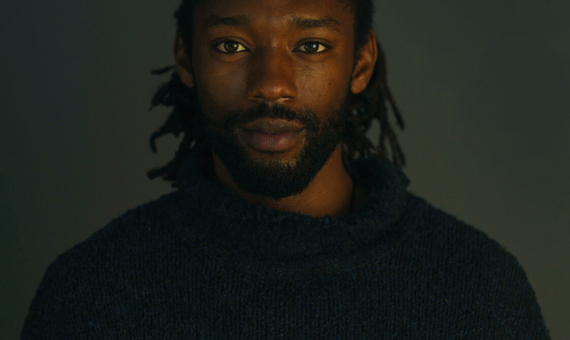 A headshot photo of Akeim Toussaint Buck. He is of Afro-Jamaican heritage with a short beard and dreadlocks tied back. He is wearing a dark blue wooly jumper and looking directly at the camera with a soft smile on his face.