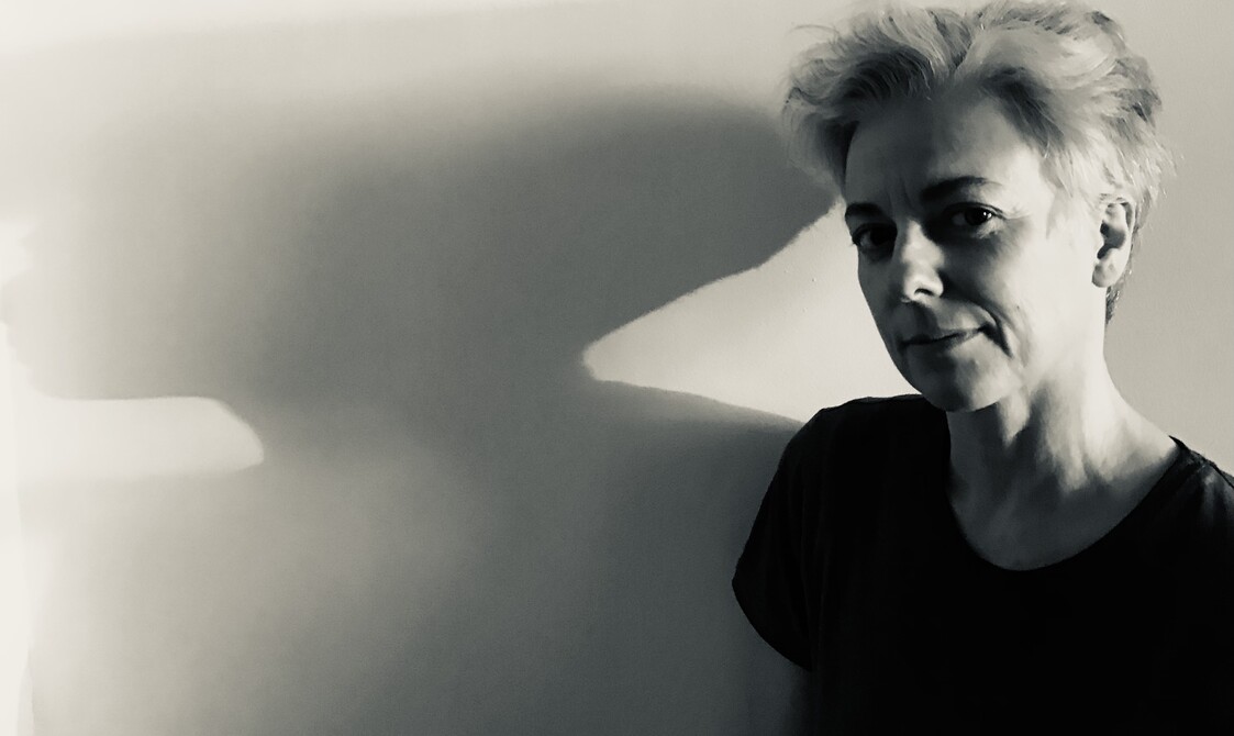 : A black and white image of Valentina Formenti, a white woman in her 50's standing against a white wall. She has short white hair and dark brown eyes. She is wearing a black short sleeve t-shirt.