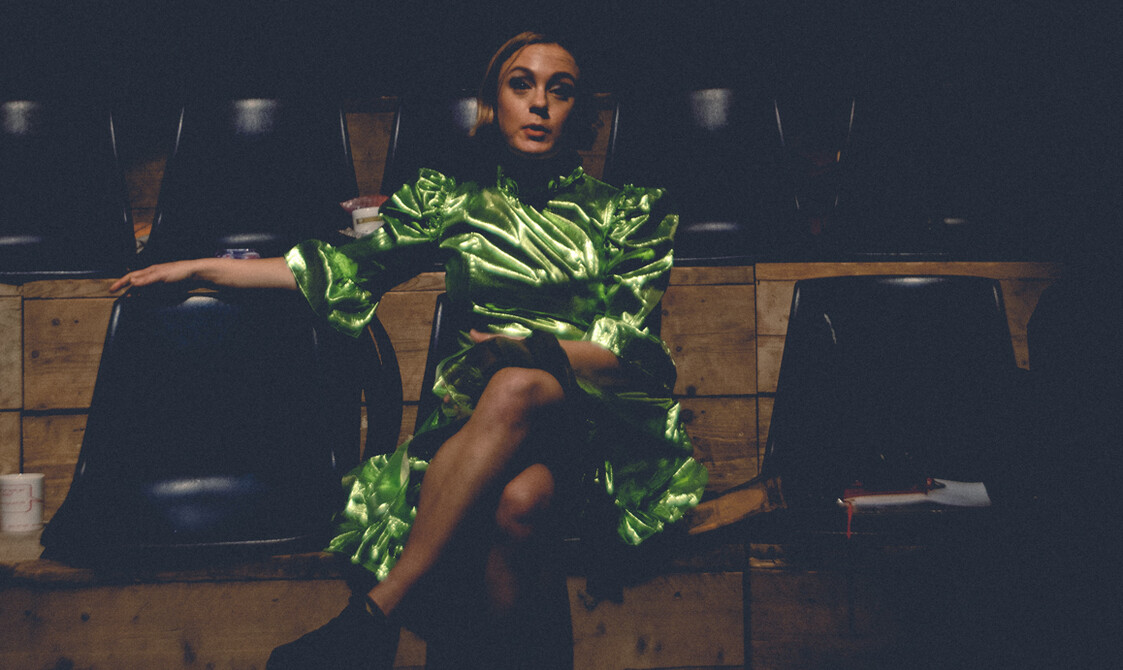 A color photo of Jessica Latowicki, a white woman in her late 30s, sitting in an empty, dark auditorium with a mug of tea and a note book to her left and right. She is speaking with her left leg crossed over her right and her right arm on the back of the seat next to her. She is wearing a bright green lamé dress with a high collar and puffy sleeves and black trainers with bare legs. She has chin length brown hair and has lots of blue eye shadow on.