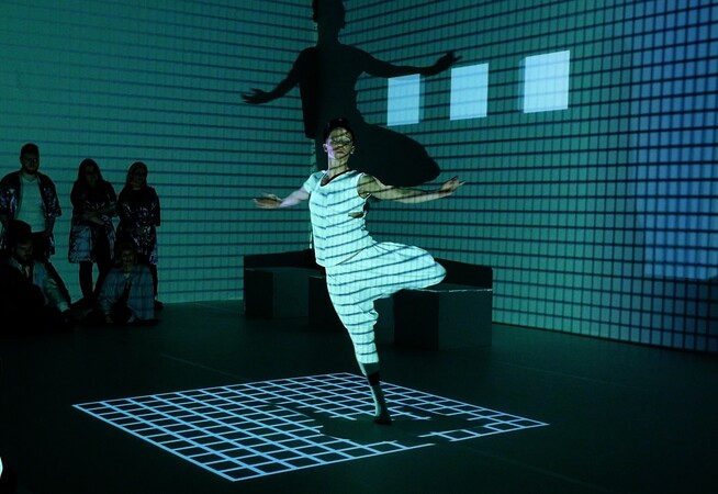 A dancer in a white vest and trousers pirouettes in a dark room, a grid of light projected onto him and the walls behind him. Audience members can be seen around the frame of the image.