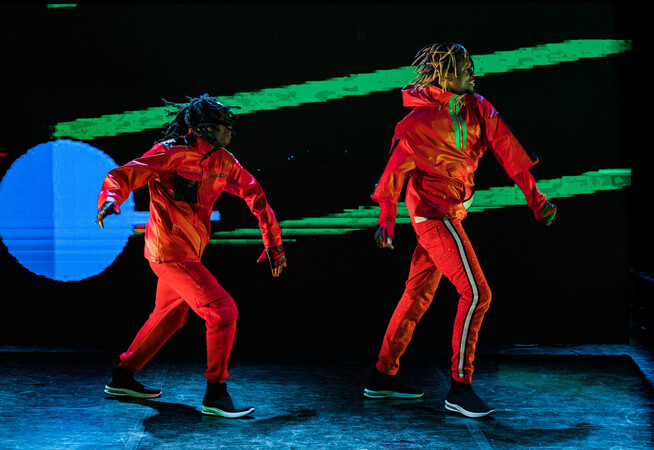 Image of two dancers in the frame. They both are wearing red tracksuits and black trainers. In the background behind them is a blue circle to the left. Two green lines also appear in the background.
