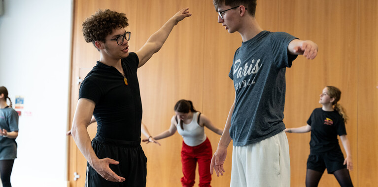 Two young students wearing glasses and dance clothing are working together. They have their arms outstretched to the side and are speaking to each other. In the background there are three other dancers