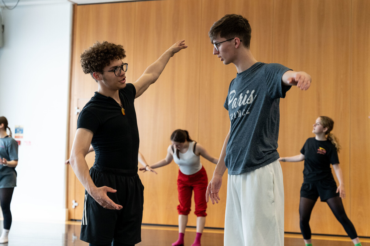 Two young students wearing glasses and dance clothing are working together. They have their arms outstretched to the side and are speaking to each other. In the background there are three other dancers