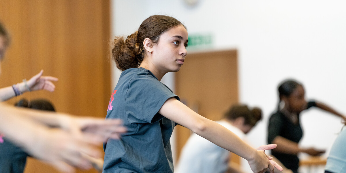 A dancer with brown hair, wearing a grey t-shirt has their arms in a curved position in front of their chest. They are looking forwards towards the mirrors in the studio