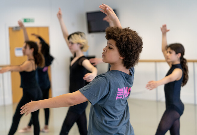 A dancer is wearing a grey t-shirt with The Place logo on the back. They are in a traditional ballet position with one arm reaching to the ceiling and the other outstretched to the side. There head is pointing diagonally as they look towards the arm to the side.