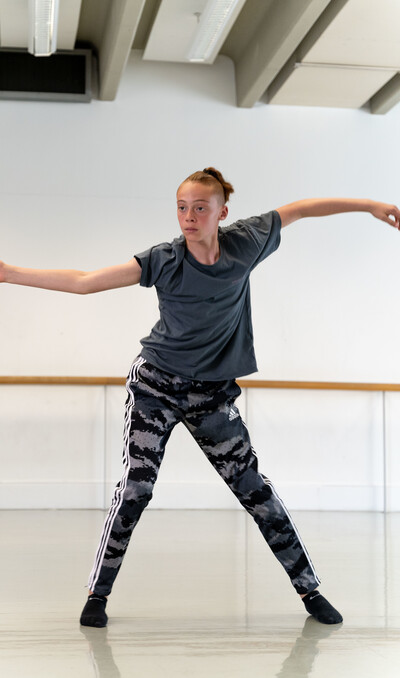 a dancer wearing a grey t-shirt and patterned grey and black trousers has one leg bent slightly and the other is stretched to the side. Their arms are curved in a wave like position out to the side and they are looking to the corner of the dance studio.