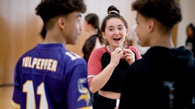 Two male students have their backs to the camera, looking at each other. One is wearing a purple sports t-shirt, the other a black hoodie. We see a young lady laughing between them. She is facing the camera and holding her necklace as she is looking at the young man on the left of the photo.